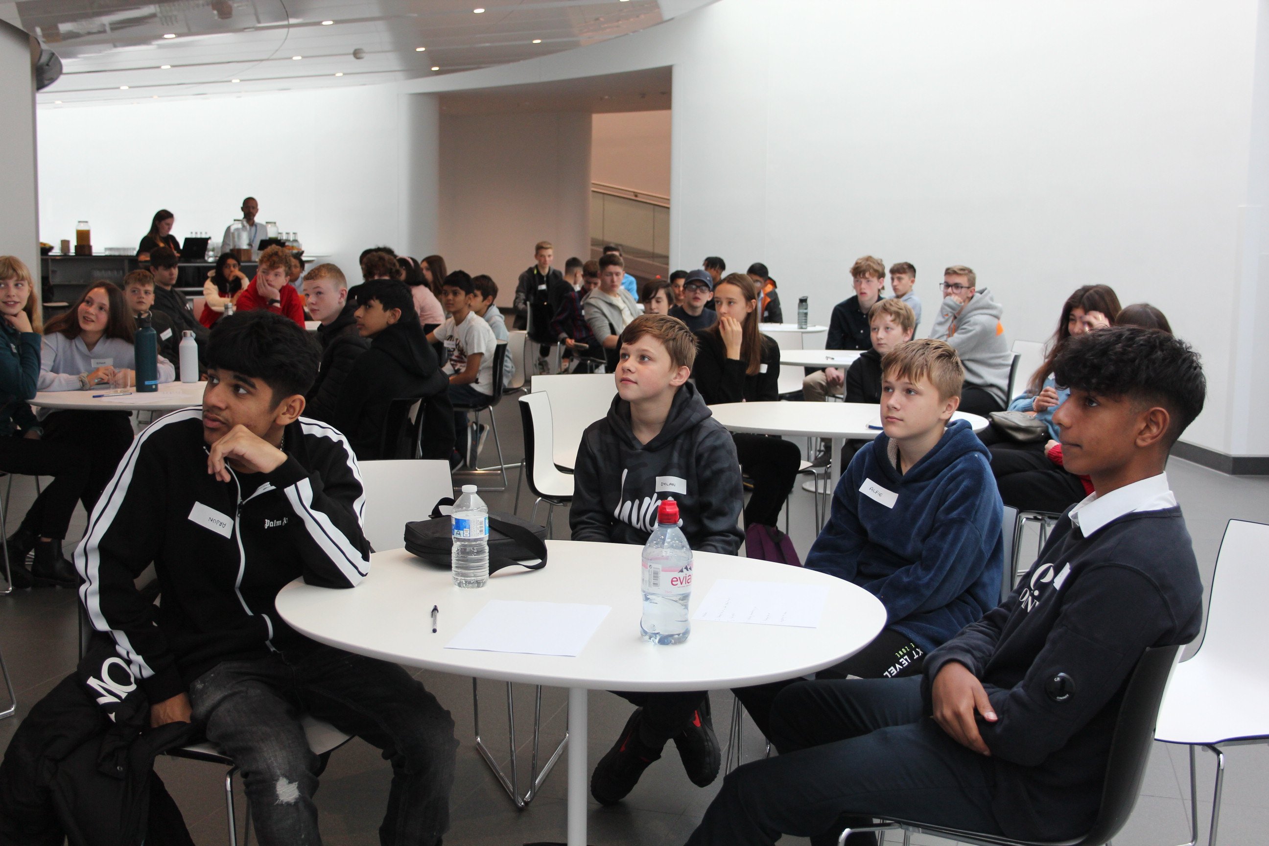 A wide shot of a group of young people sitting at round white tables in a large, modern room with white walls and high ceilings. They are facing a person who is not visible in the shot, likely listening to a presentation. 