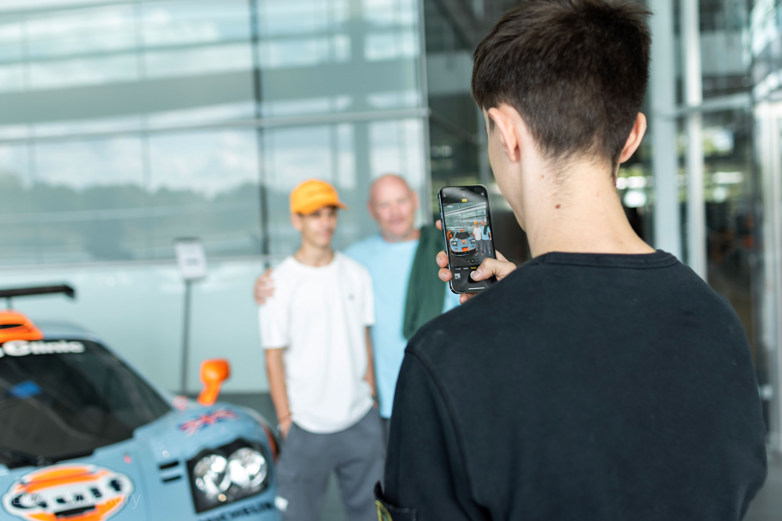 A person is holding a smartphone, taking a picture of two people standing in front of a McLaren race car.