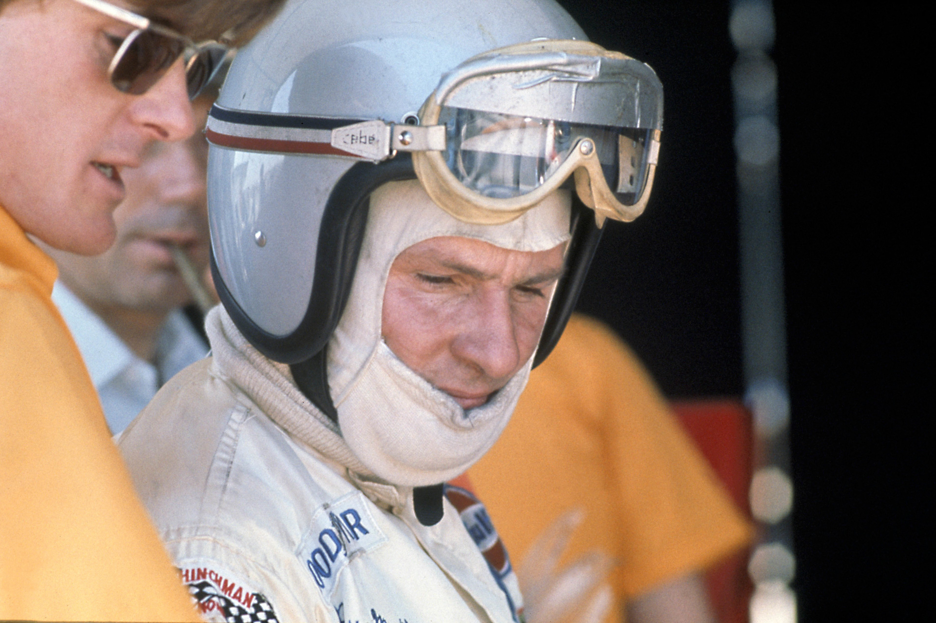 A close-up of racing driver Bruce McLaren wearing a helmet and goggles.