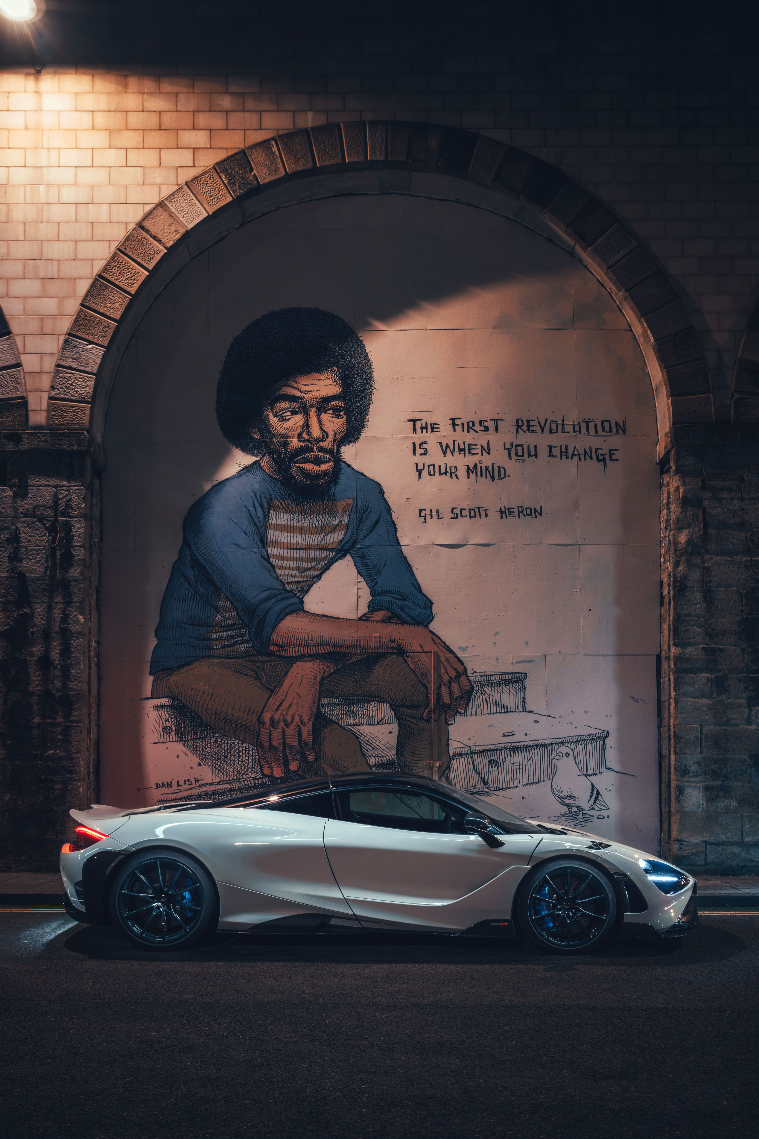 A white McLaren supercar parked at night in front of a Gil Scott-Heron mural.