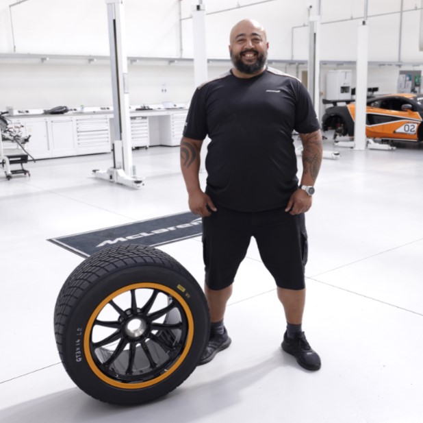 A McLaren employee smiling, standing in a McLaren facility next to a wheel.