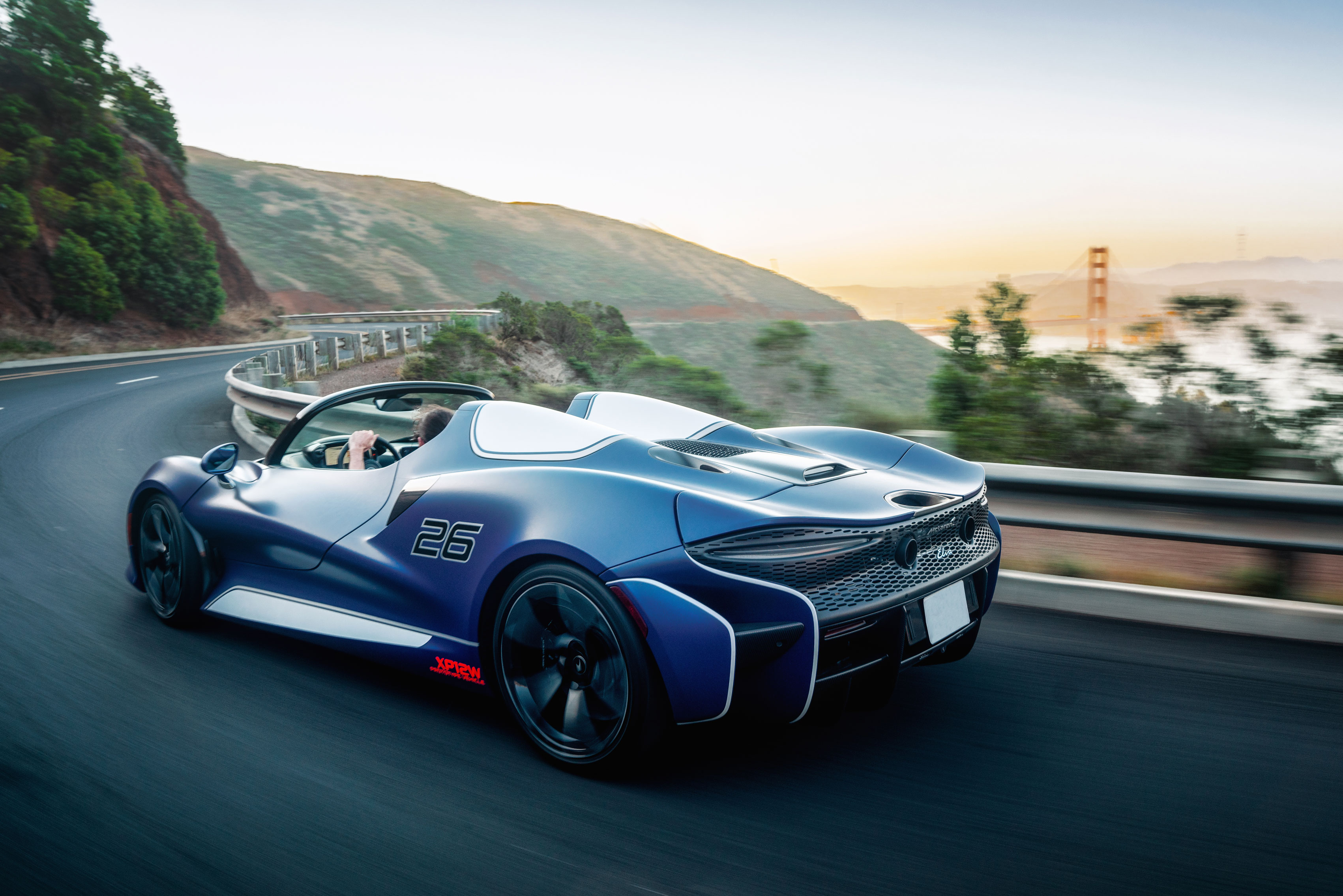  A blue and white McLaren Elva supercar driving on a winding road with the Golden Gate Bridge in the distance.