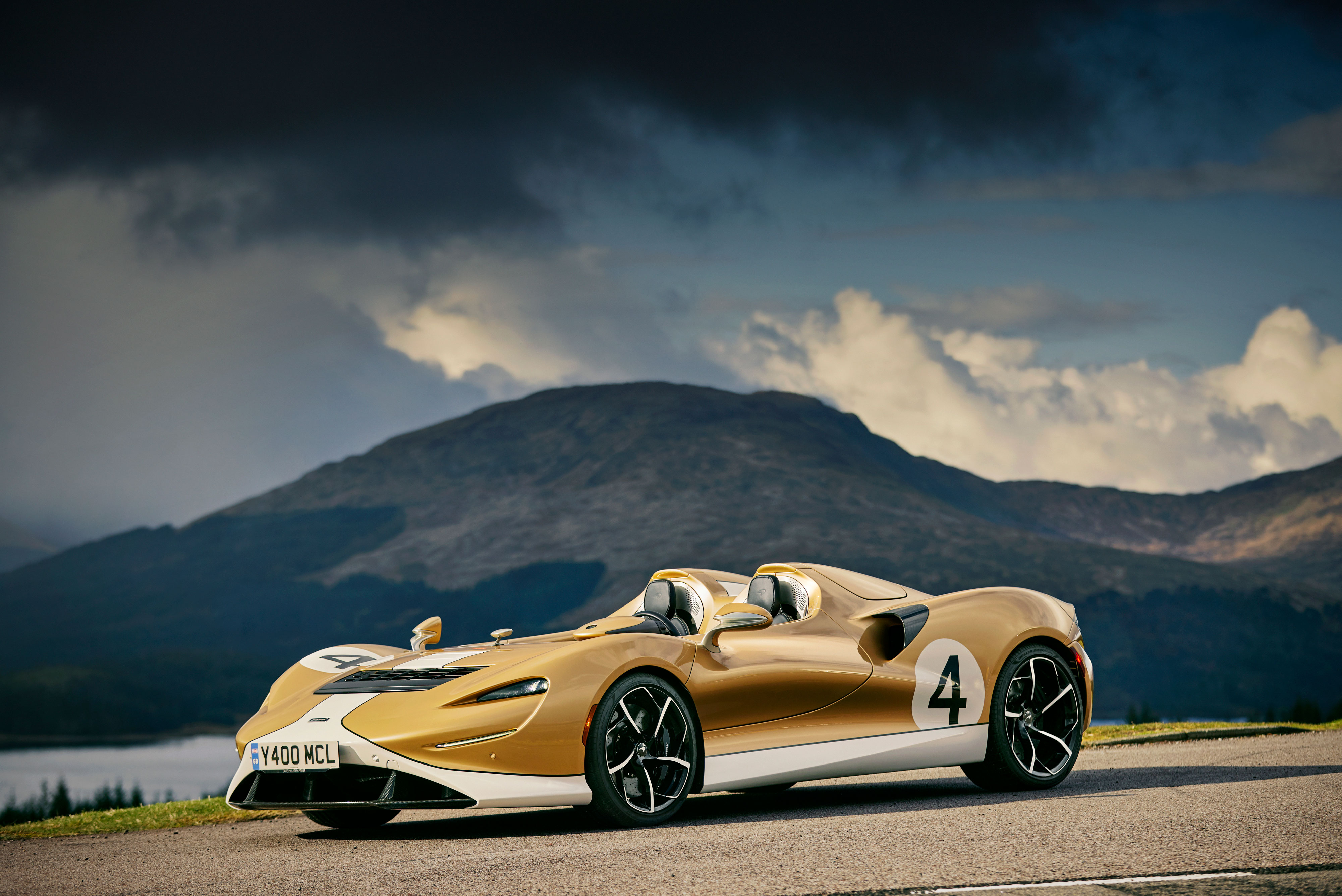 A gold and white McLaren Elva supercar with a racing number, parked outdoors with a mountain backdrop.
