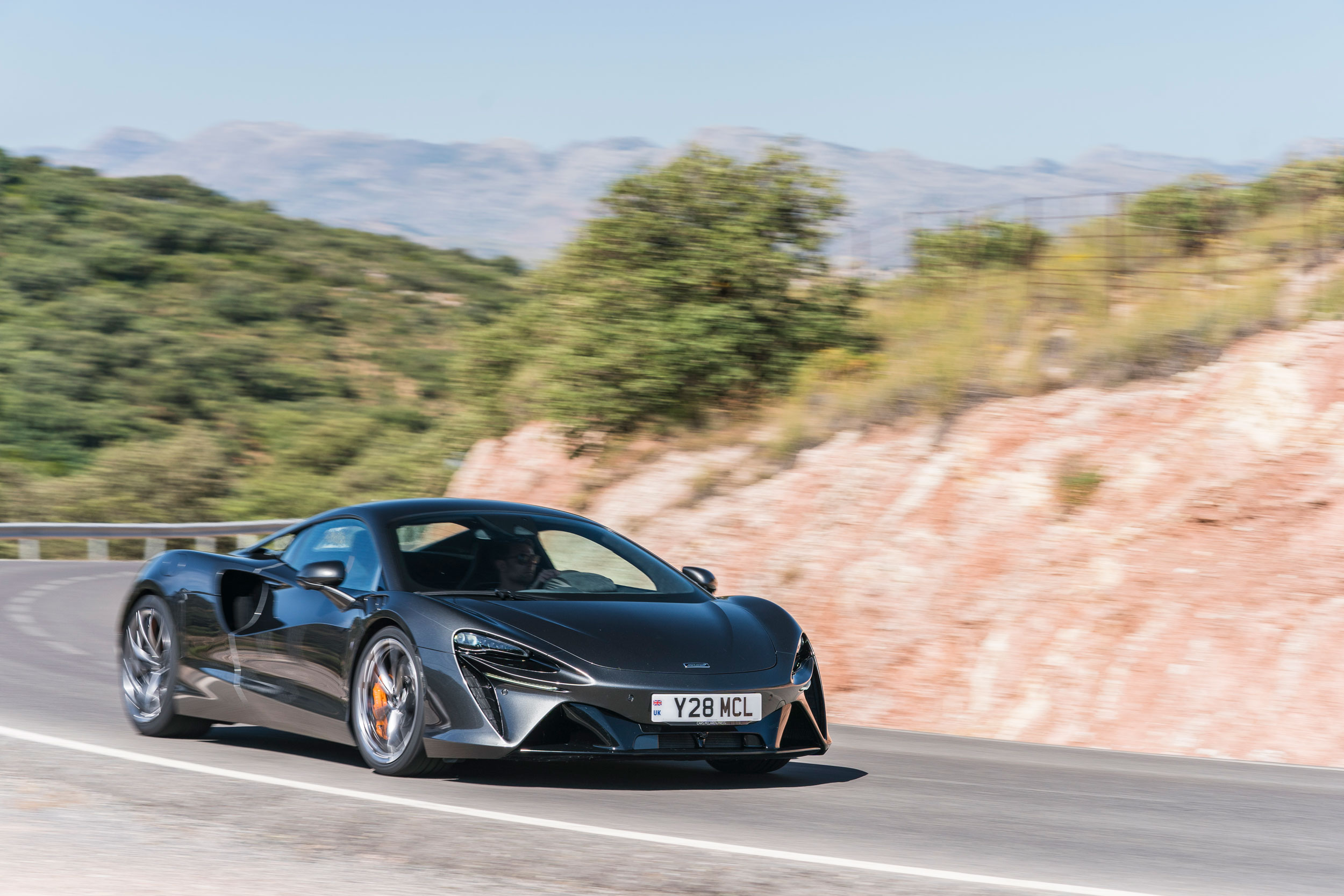 A dark grey McLaren Artura supercar driving on a winding road with a mountain background.