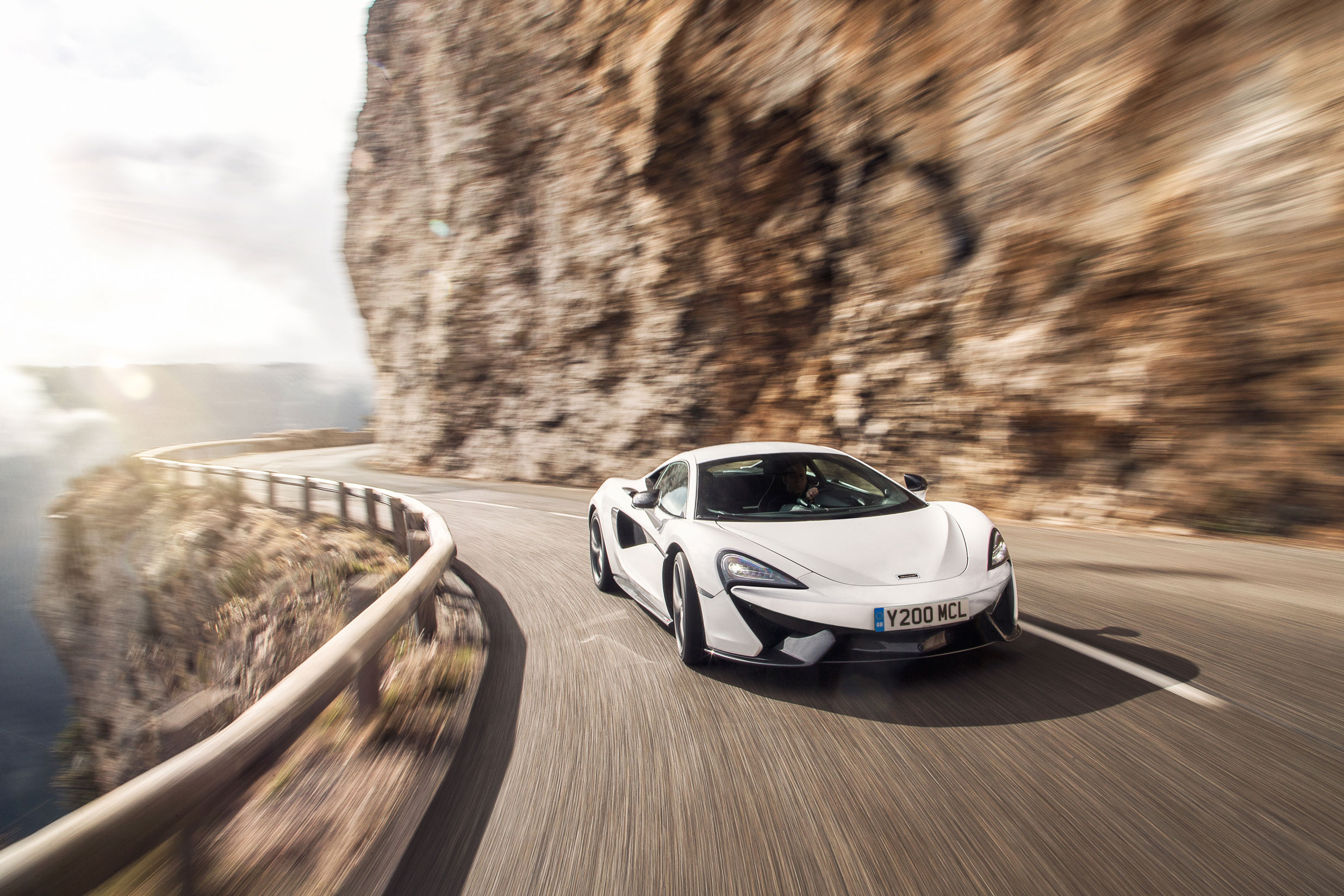 A white McLaren 570S supercar driving fast on a winding mountain road.