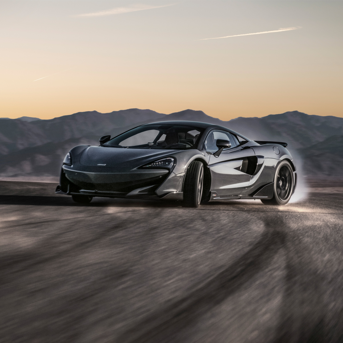 A dark grey McLaren 570S supercar drifting in a desert landscape at sunset.