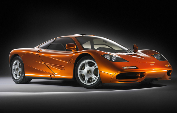A front-side view of an orange McLaren F1 supercar, shot in a dark studio.