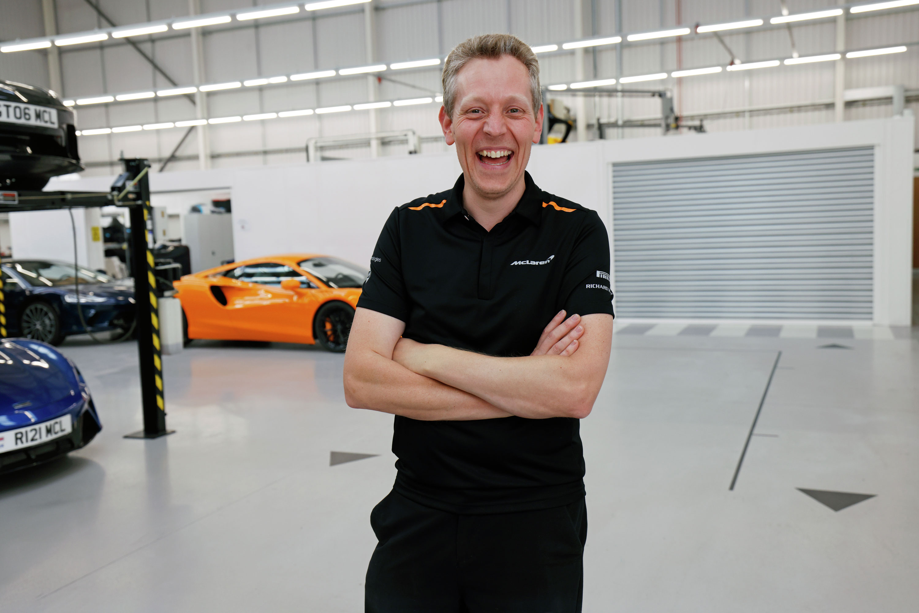  A laughing man in a black McLaren polo shirt, with his arms crossed, standing in a workshop. An orange McLaren Artura is in the background.