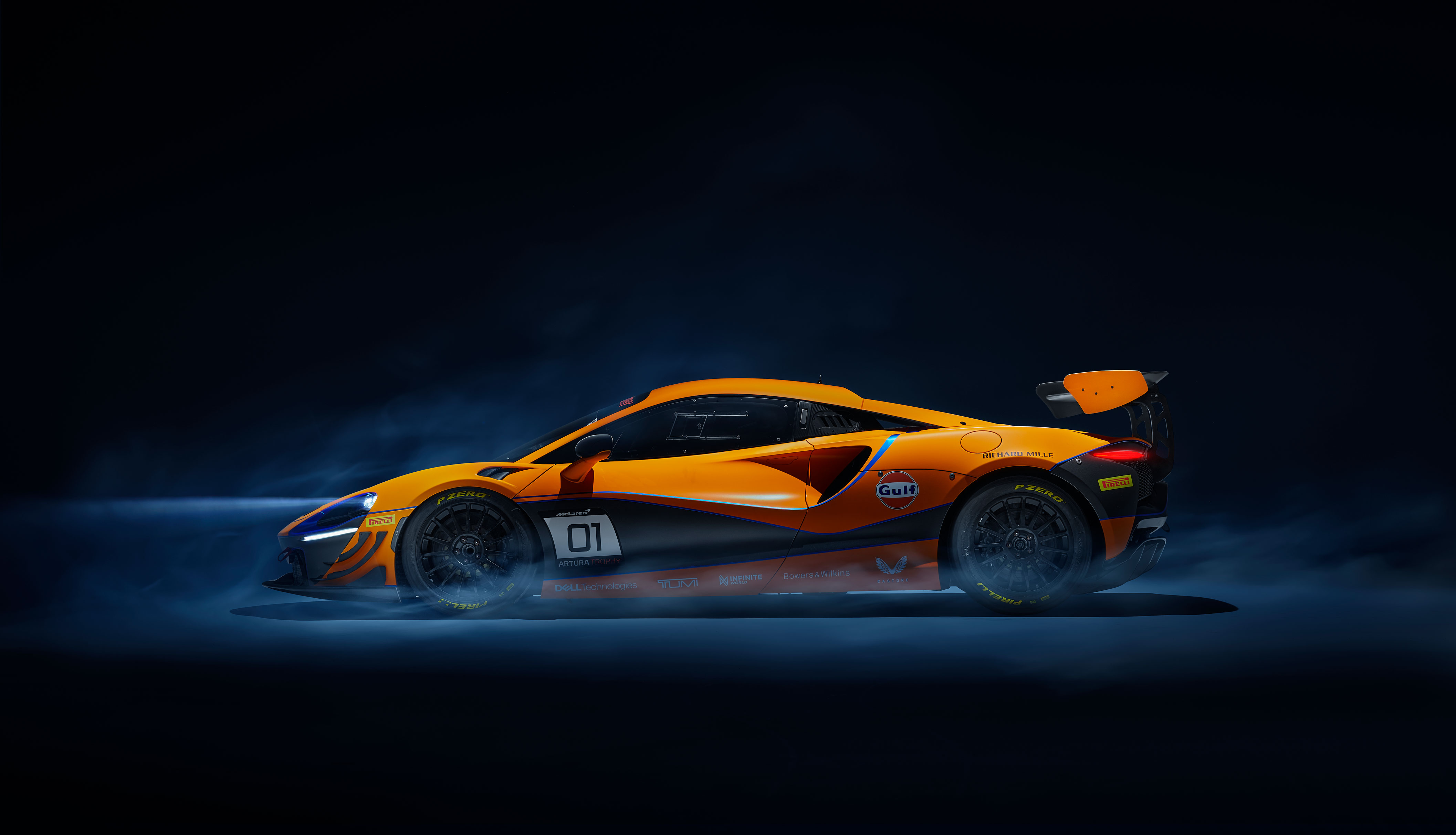 An orange McLaren Artura GT4 race car with Gulf livery, in a dark, smoky setting.
