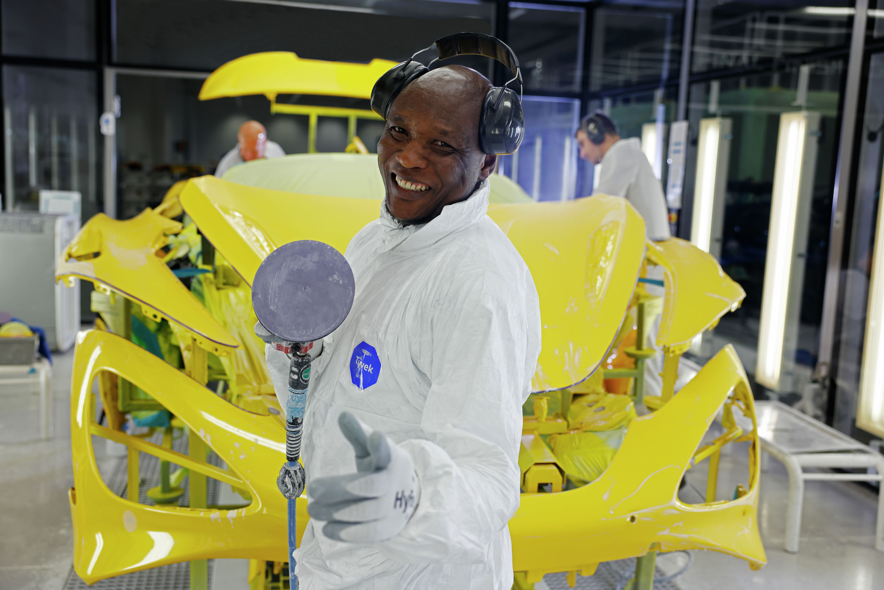 A happy McLaren factory worker in a white jumpsuit, holding a tool and standing next to a yellow car body.