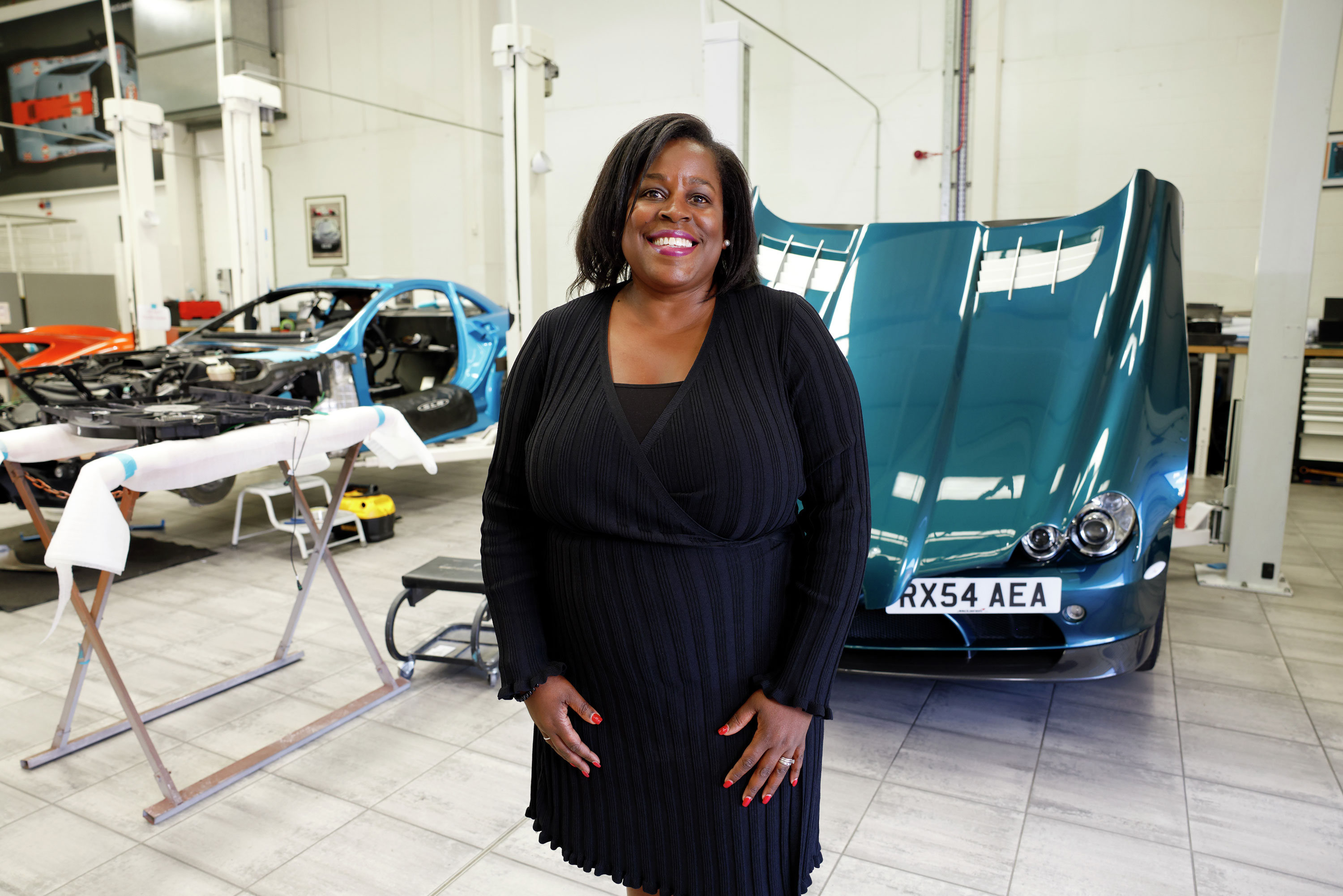 A smiling woman standing in a McLaren workshop with a teal car in the background.