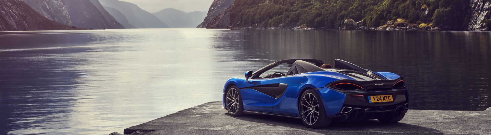 McLaren 570S Spider parked at the water's edge, highlighting the car's sleek design and the stunning natural landscape.