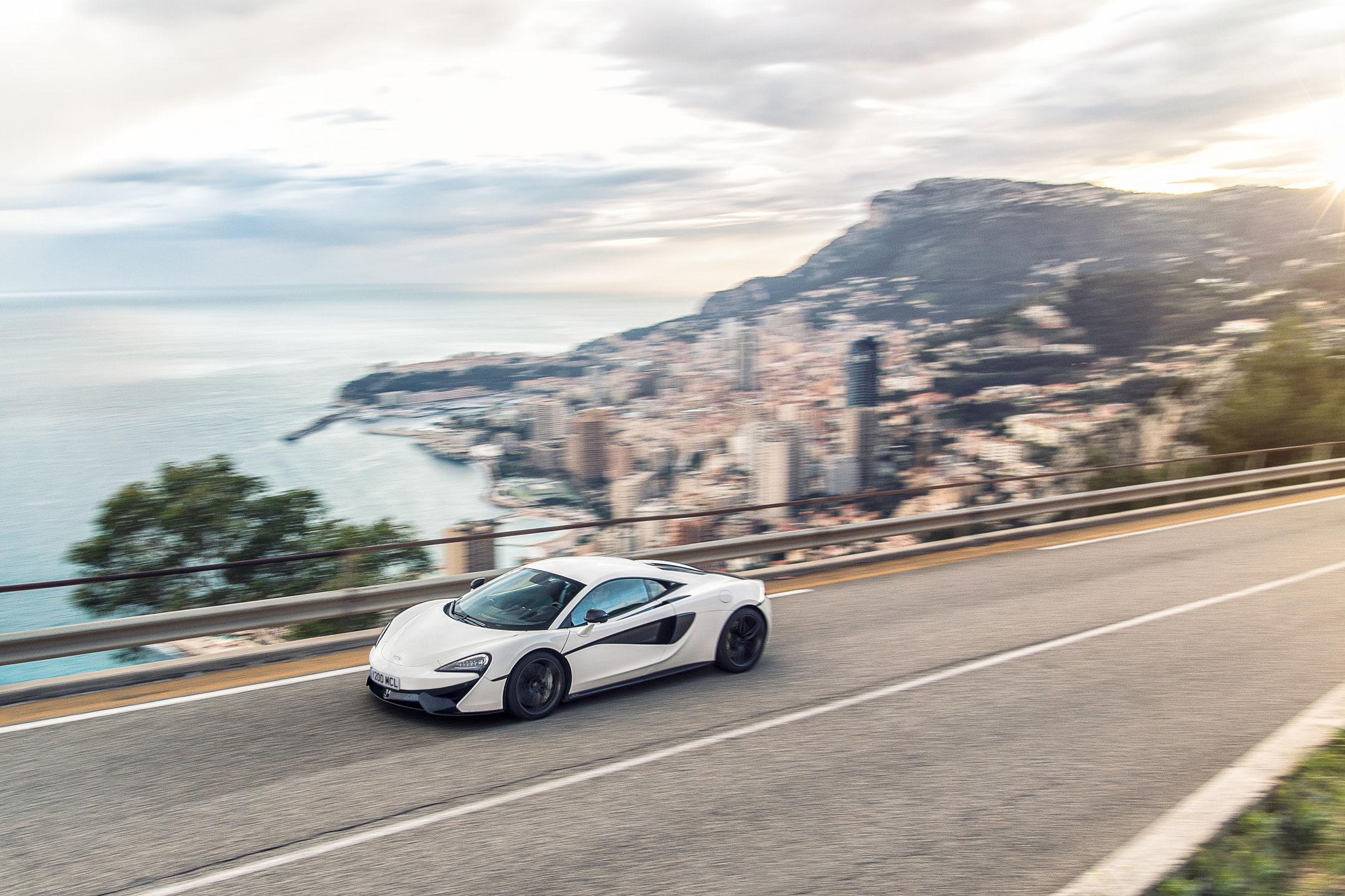 A white McLaren 570S supercar driving on a winding road with a blurred city and coastline in the background.